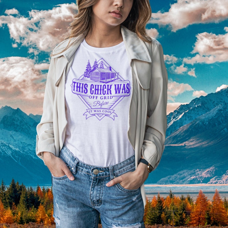 May include: A white t-shirt featuring a purple graphic design of a cabin and the text "This Chick Was Off Grid Before It Was Cool". The person wearing the shirt is standing in front of a mountain landscape.