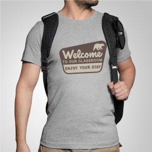 Puede incluir: Una camiseta gris jaspeada con un letrero vintage marr&oacute;n y blanco que dice "Bienvenido a nuestra aula, disfruta de tu estancia". Un oso de dibujos animados camina a la derecha del letrero.