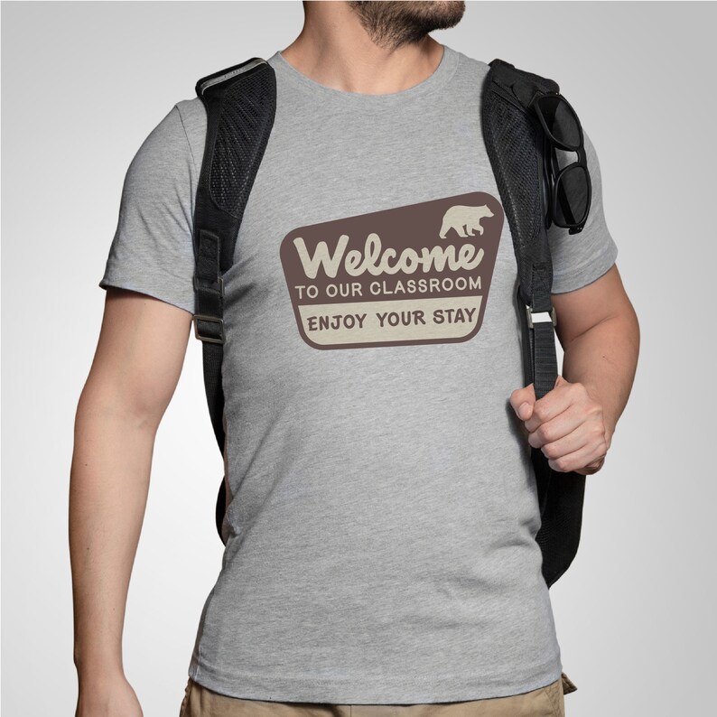 Puede incluir: Una camiseta gris jaspeada con un letrero vintage marr&oacute;n y blanco que dice "Bienvenido a nuestra aula, disfruta de tu estancia". Un oso de dibujos animados camina a la derecha del letrero.