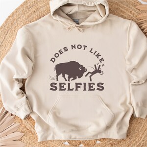 May include: A beige hoodie with a brown graphic of a buffalo chasing a stick figure. The text on the hoodie reads "Does Not Like Selfies".