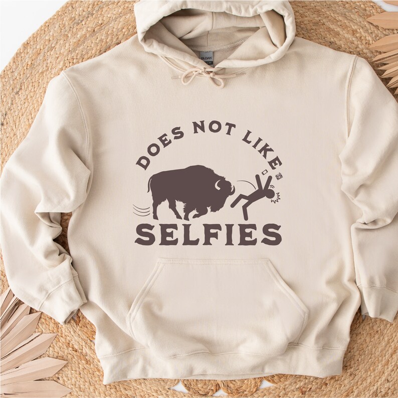 May include: A beige hoodie with a brown graphic of a buffalo chasing a stick figure. The text on the hoodie reads "Does Not Like Selfies".