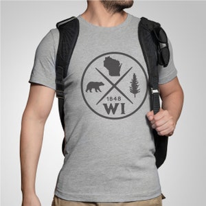 Pode incluir: Uma camiseta cinza com um design gr&aacute;fico preto e branco que apresenta um urso, um pinheiro, o contorno do Wisconsin e o texto "1848 WI".