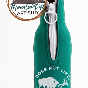 May include: Green zippered bottle cooler with a white buffalo and a stick figure running away from it. The text reads "Does Not Like Selfies".
