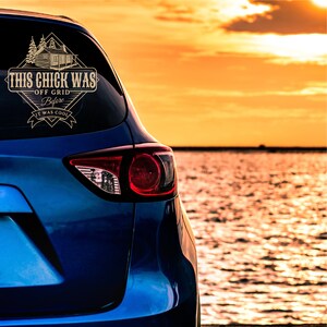 May include: A blue car with a gold decal on the rear window that reads "This Chick Was Off Grid Before It Was Cool". The car is parked by a body of water with a sunset in the background.