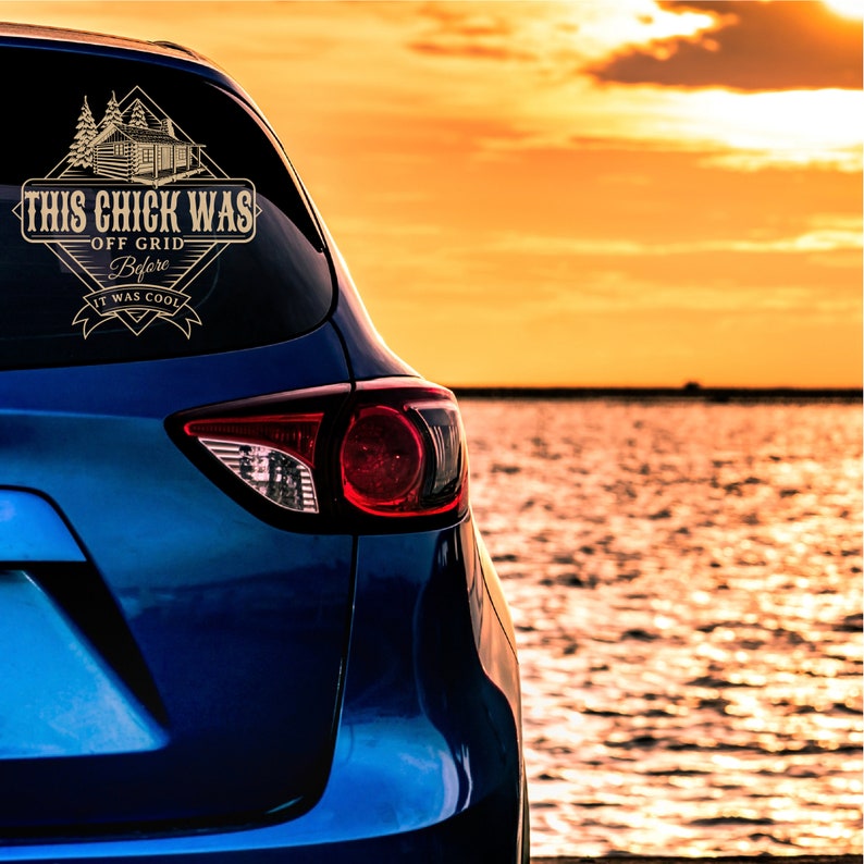 May include: A blue car with a gold decal on the rear window that reads "This Chick Was Off Grid Before It Was Cool". The car is parked by a body of water with a sunset in the background.