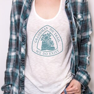 May include: White tank top with a blue graphic of a hiking boot with sunflowers and the text "Grandmas on Trail We Do Exist".