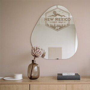 May include: A brown, irregularly shaped mirror with a brown graphic of a mountain range, trees, and a bear with the text "NEW MEXICO - EST. 1912 -" inside a hexagon. A pink protea flower in a clear glass vase sits on a wooden surface in front of the mirror.
