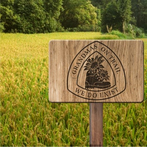 May include: A wooden sign with a brown background and a white circle with the text "Grandmas on Trail We Do Exist" in black lettering. The circle has a stylized image of a hiking boot with sunflowers.