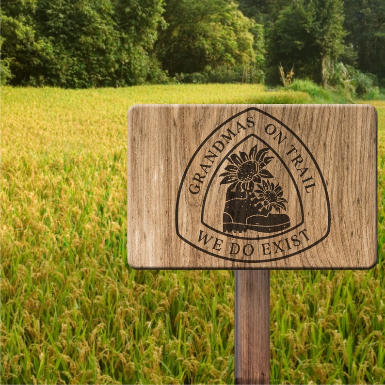May include: A wooden sign with a brown background and a white circle with the text "Grandmas on Trail We Do Exist" in black lettering. The circle has a stylized image of a hiking boot with sunflowers.
