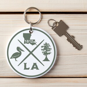 May include: White round keychain with a green pelican, tree, and the state of Louisiana. The keychain also has the text "LA" and "1812" on it.