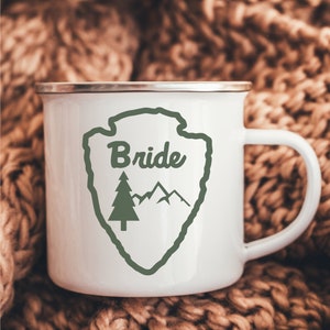 May include: White enamel mug with a green and white design of a mountain range and a pine tree. The design is inside a green outline of a shield shape. The text "Bride" is written in cursive inside the shield.
