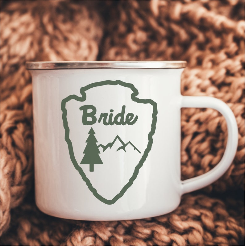 May include: White enamel mug with a green and white design of a mountain range and a pine tree. The design is inside a green outline of a shield shape. The text "Bride" is written in cursive inside the shield.