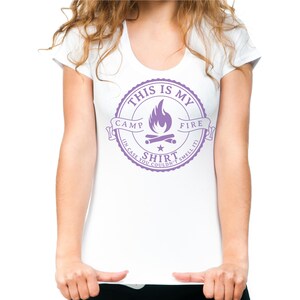 May include: White scoop-neck t-shirt with a purple graphic. The graphic reads "THIS IS MY CAMP FIRE SHIRT (IN CASE YOU COULDN'T SMELL IT)" and features a campfire design. The shirt is a casual wear item.