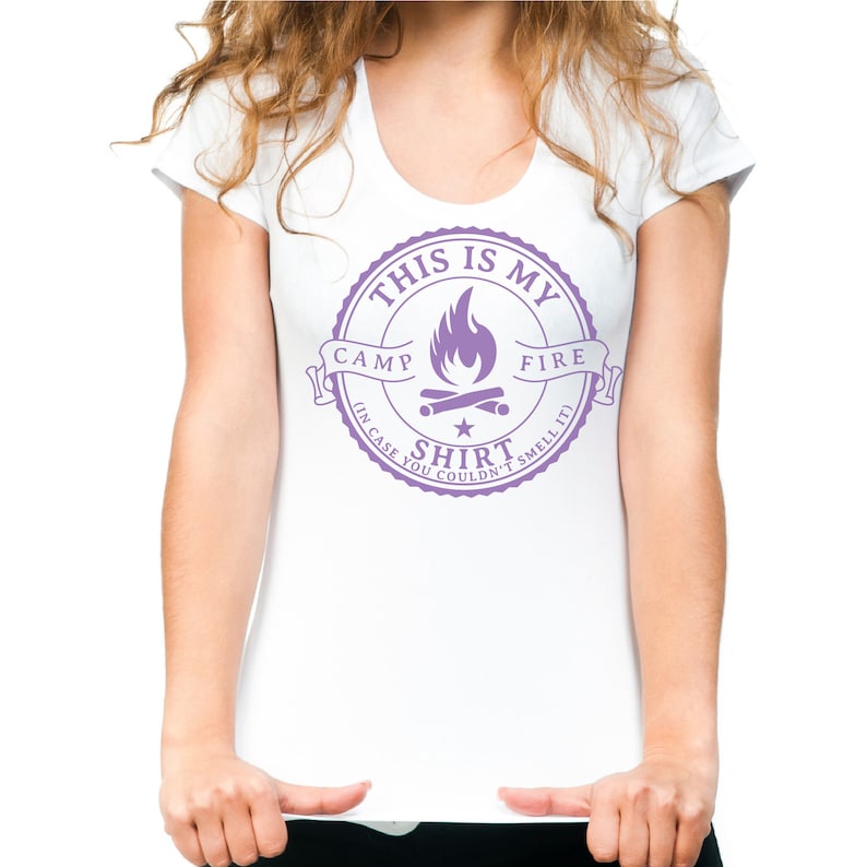 May include: White scoop-neck t-shirt with a purple graphic. The graphic reads "THIS IS MY CAMP FIRE SHIRT (IN CASE YOU COULDN'T SMELL IT)" and features a campfire design. The shirt is a casual wear item.