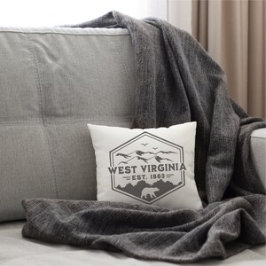 May include: White throw pillow with a gray graphic design of a mountain range and a bear. The graphic includes the text "West Virginia - Est. 1863 - ". The pillow is on a gray couch with a gray throw blanket.