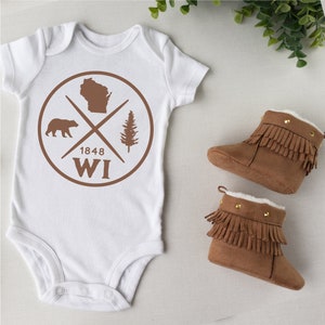Pode incluir: Macac&atilde;o branco para beb&ecirc; com um gr&aacute;fico circular marrom que apresenta um urso, um pinheiro, o estado de Wisconsin e o texto "1848 WI". Duas botinhas marrons para beb&ecirc; com franjas e detalhes dourados.