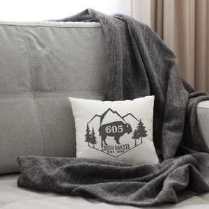 May include: White throw pillow with a black and white graphic design featuring a mountain range, a silhouette of a bison, and the text "605 South Dakota Est. 1889".