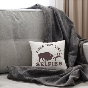 May include: White throw pillow with brown text that reads "DOES NOT LIKE SELFIES". A brown silhouette of a buffalo is shown charging at a stick figure holding a phone.