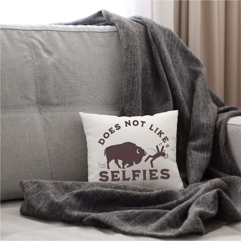 May include: White throw pillow with brown text that reads "DOES NOT LIKE SELFIES". A brown silhouette of a buffalo is shown charging at a stick figure holding a phone.