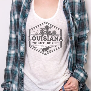 May include: White tank top with a grey graphic design of a hexagon with a tree, a bear, and the text "Louisiana Est. 1812".