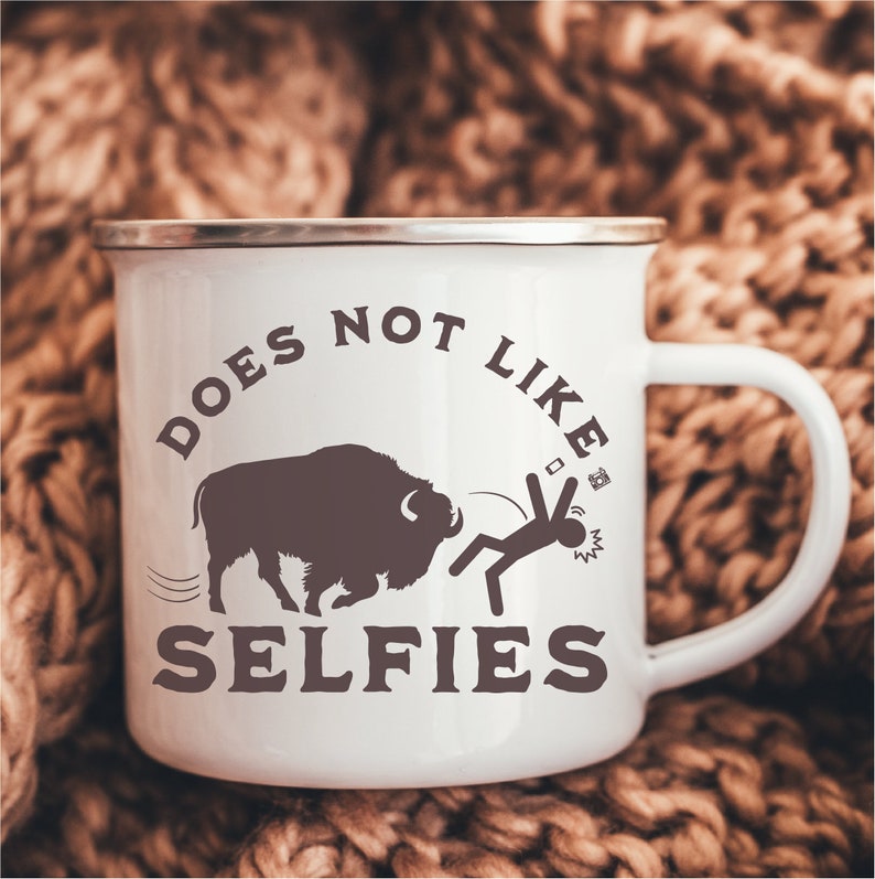 May include: White enamel mug with a brown silhouette of a buffalo and a stick figure running away from it. The text on the mug reads "Does not like selfies".