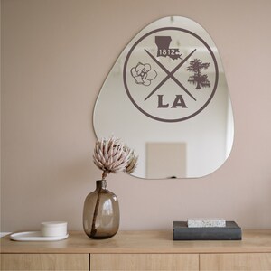 May include: A round, irregular-shaped mirror with a brown graphic of a magnolia flower, a cypress tree, and the state of Louisiana with the text "1812" and "LA" in a circle.