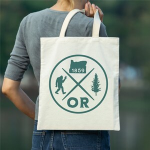 May include: A white canvas tote bag with a green circle design. The circle features a silhouette of the state of Oregon, a pine tree, a Bigfoot, and the text "OR" and "1859".