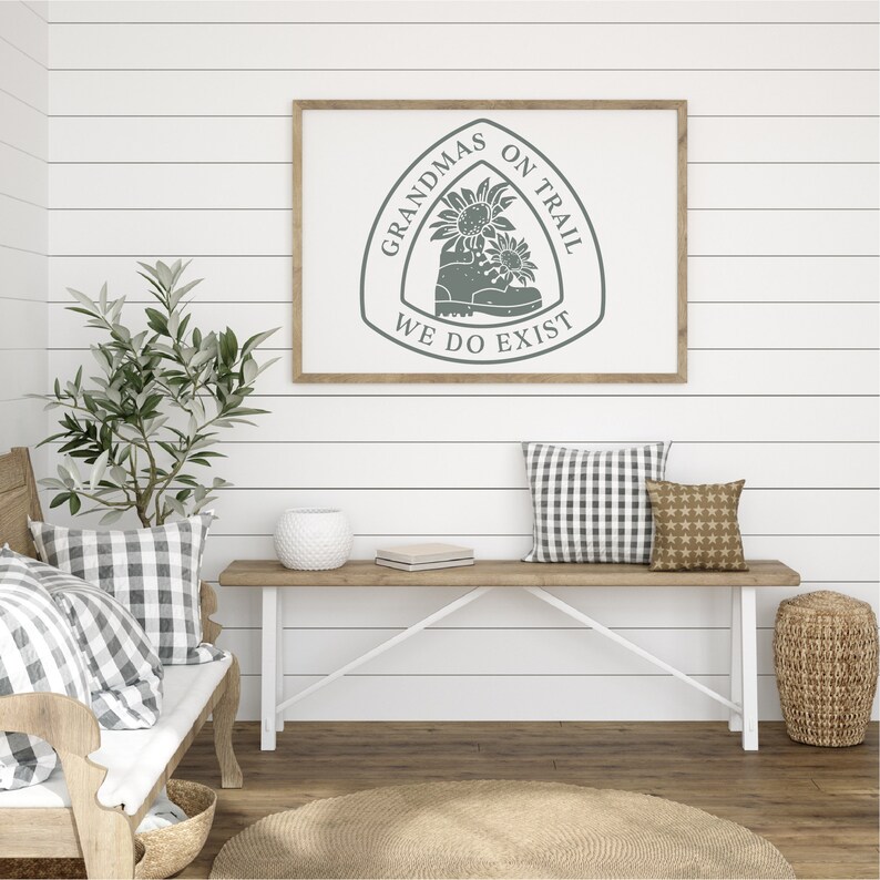 May include: A framed print with a gray silhouette of a hiking boot with sunflowers on top. The text around the boot reads "Grandmas on Trail We Do Exist". The print is hanging on a white shiplap wall above a wooden bench with pillows.