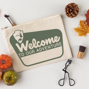 May include: A beige canvas zippered pouch with a green welcome sign that says "Welcome to our adventure". The sign has a brown silhouette of a park ranger badge with a pine tree and a bear inside.