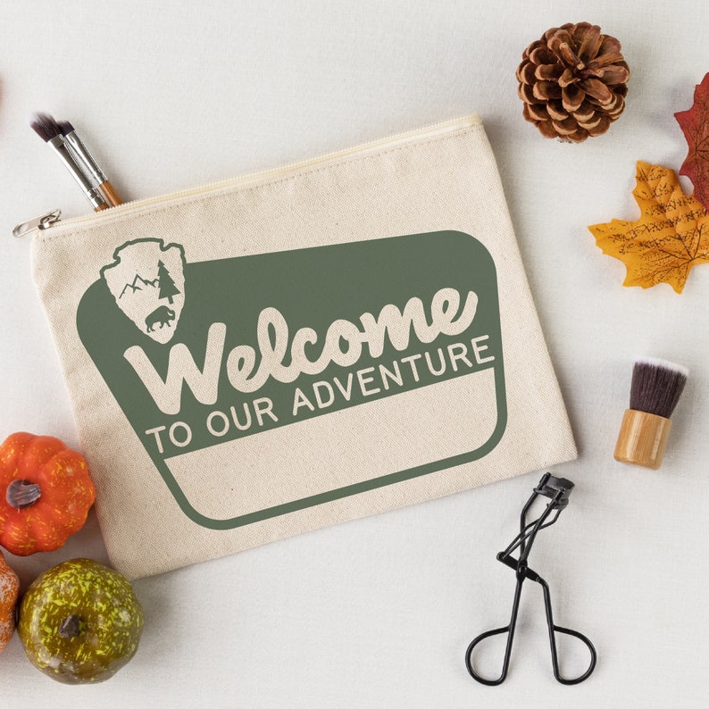 May include: A beige canvas zippered pouch with a green welcome sign that says "Welcome to our adventure". The sign has a brown silhouette of a park ranger badge with a pine tree and a bear inside.