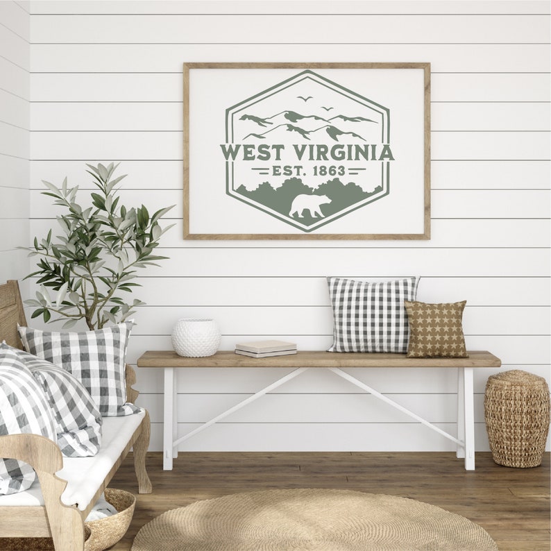 May include: A framed print with a green mountain and bear design. The print says "West Virginia - Est. 1863". The print is hanging on a white shiplap wall above a wooden bench.