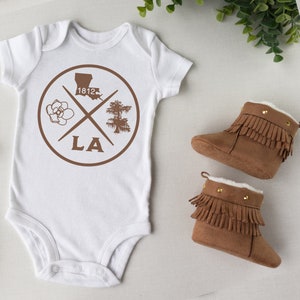 May include: White baby onesie with a brown circle design featuring a magnolia flower, a cypress tree, the state of Louisiana, and the text "LA" and "1812". Two brown suede baby booties with fringe and gold accents.