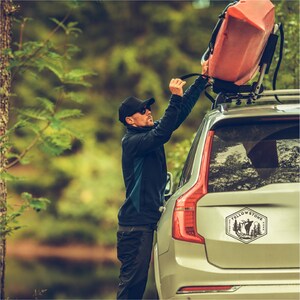 May include: A person is securing a red kayak to the roof rack of a silver SUV. The SUV has a "Yellowstone" sticker on the back window with a silhouette of an elk and pine trees.