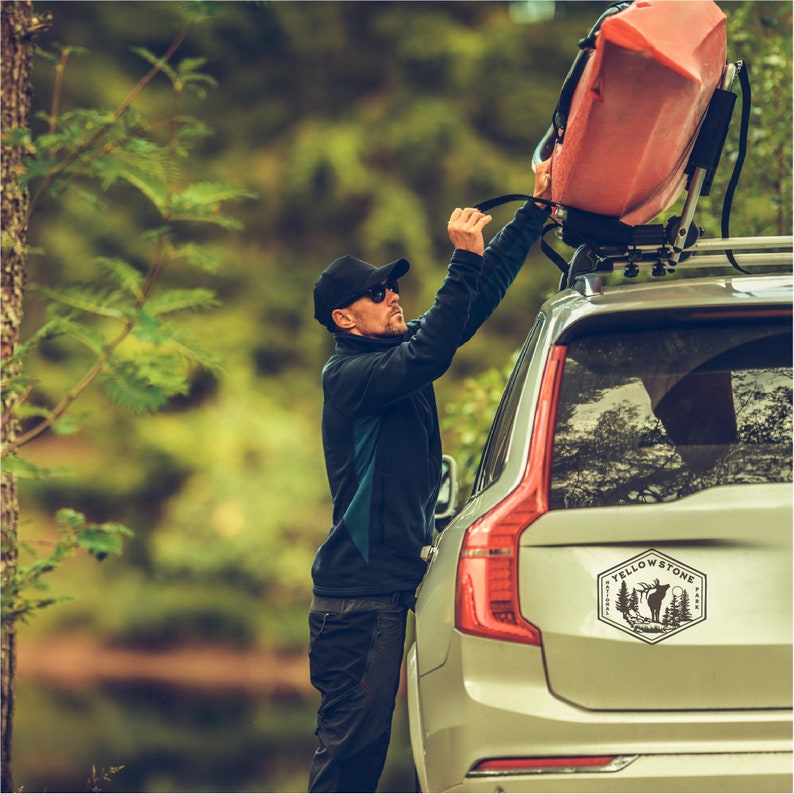 May include: A person is securing a red kayak to the roof rack of a silver SUV. The SUV has a "Yellowstone" sticker on the back window with a silhouette of an elk and pine trees.