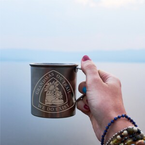 May include: A silver camping mug with a black and white graphic of a hiking boot and the text "Grandmas on Trail We Do Exist".