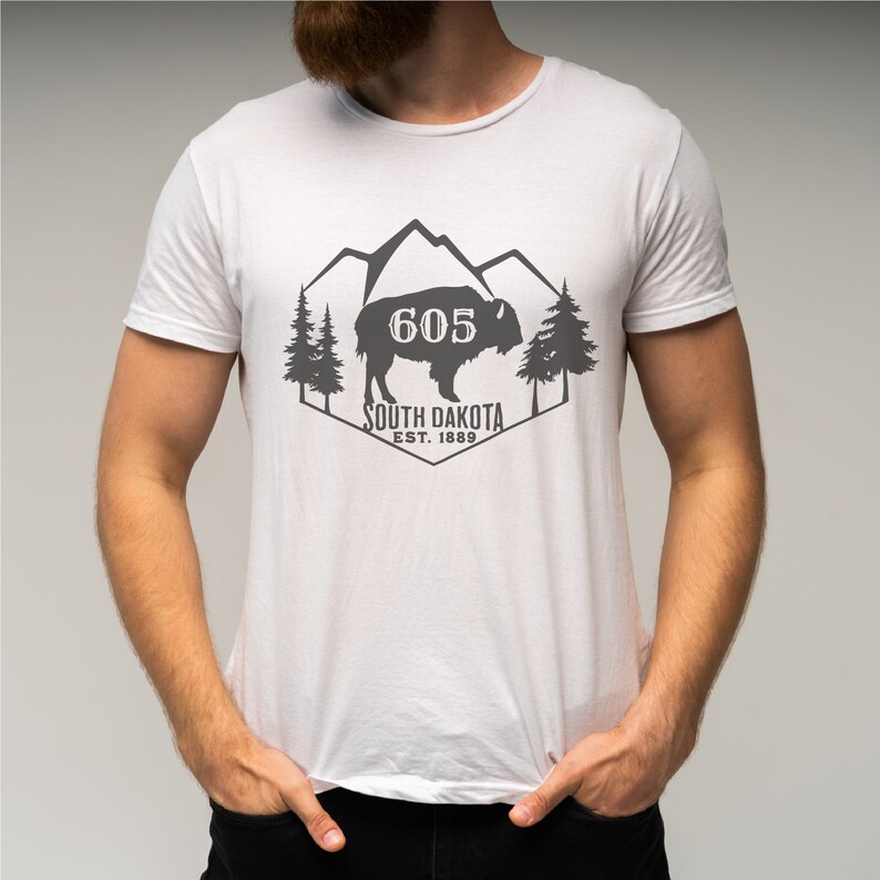 May include: White t-shirt with a gray graphic design featuring a silhouette of a bison in front of mountains and trees. The text "605 South Dakota Est. 1889" is printed below the bison.