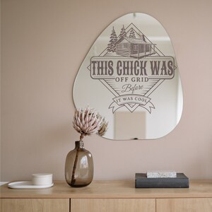 May include: A decorative mirror with a rustic cabin design and the text "This Chick Was Off Grid Before It Was Cool". The mirror is shaped like a teardrop and has a brown wooden frame.