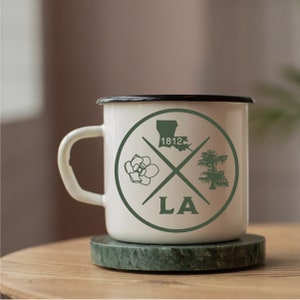 May include: White enamel mug with a black rim and a green design. The design features a magnolia flower, a cypress tree, the state of Louisiana, and the text "LA" and "1812".