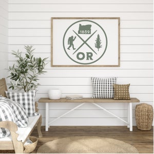 May include: A framed print with a green circle design featuring a silhouette of the state of Oregon, a Bigfoot, a pine tree, and the text "1859 OR". The print is hanging on a white shiplap wall.