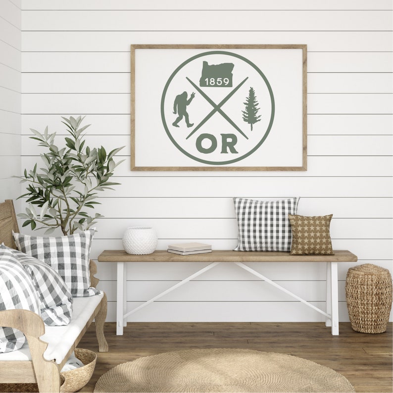 May include: A framed print with a green circle design featuring a silhouette of the state of Oregon, a Bigfoot, a pine tree, and the text "1859 OR". The print is hanging on a white shiplap wall.