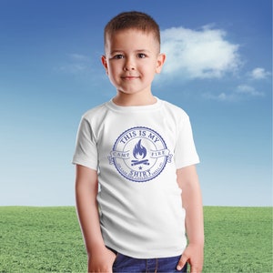 May include: A white t-shirt with a blue graphic that reads "THIS IS MY CAMP FIRE SHIRT (IN CASE YOU COULDN'T SMELL IT!)". The design features a campfire illustration inside a circular emblem. The shirt is worn by a young child.
