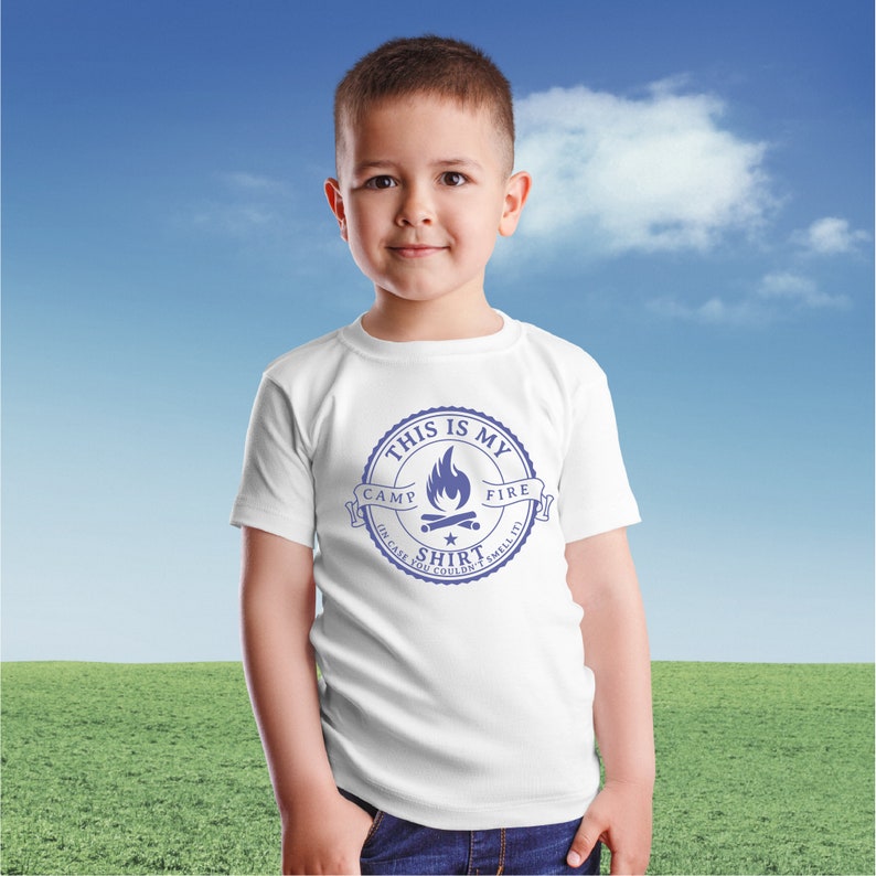 May include: A white t-shirt with a blue graphic that reads "THIS IS MY CAMP FIRE SHIRT (IN CASE YOU COULDN'T SMELL IT!)". The design features a campfire illustration inside a circular emblem. The shirt is worn by a young child.