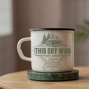 May include: White enamel mug with a black rim and a green graphic of a cabin in the woods. The graphic says "This Guy Was Off Grid Before It Was Cool".