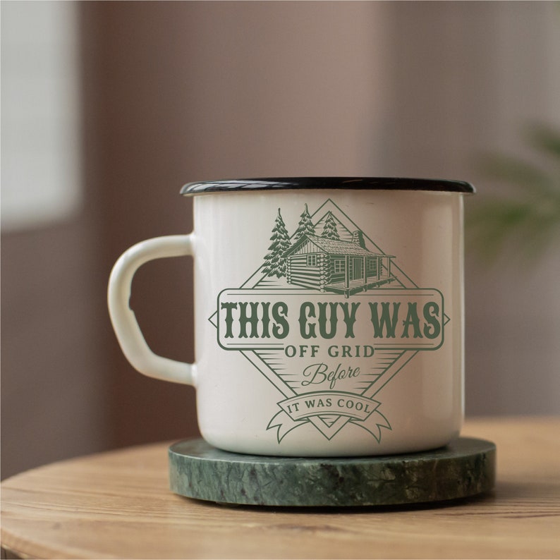 May include: White enamel mug with a black rim and a green graphic of a cabin in the woods. The graphic says "This Guy Was Off Grid Before It Was Cool".