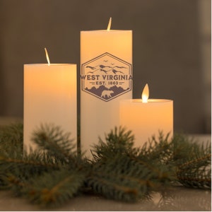 May include: Three white pillar candles with a flickering flame. The middle candle has a brown graphic of a mountain range with the text "West Virginia - Est. 1863". The candles are surrounded by green pine branches.