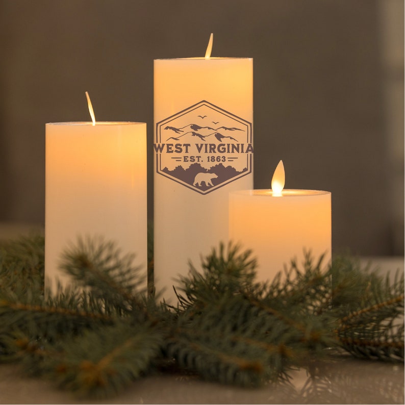 May include: Three white pillar candles with a flickering flame. The middle candle has a brown graphic of a mountain range with the text "West Virginia - Est. 1863". The candles are surrounded by green pine branches.