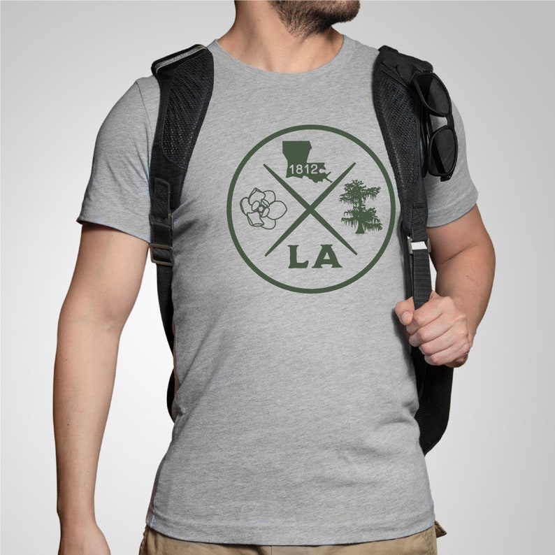 May include: A gray t-shirt with a green circle design featuring a magnolia flower, a cypress tree, the outline of Louisiana, and the text "1812" and "LA".