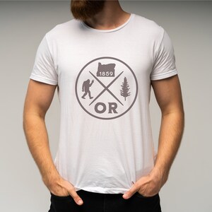 May include: White t-shirt with a grey graphic design featuring a stylised outline of the state of Oregon, a pine tree, a Bigfoot, and the text "OR" and "1859".
