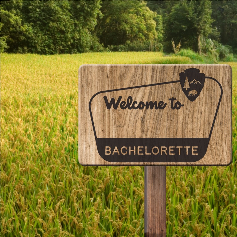 May include: A wooden sign with a brown background and a white border. The sign says "Welcome to Bachelorette" in black text. The sign is in front of a field of yellow grass.