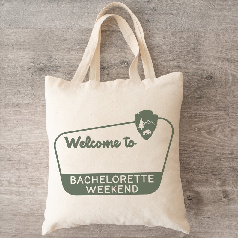 May include: A white canvas tote bag with green text that says "Welcome to Bachelorette Weekend". The bag has a green park ranger logo with a bison, trees, and mountains.